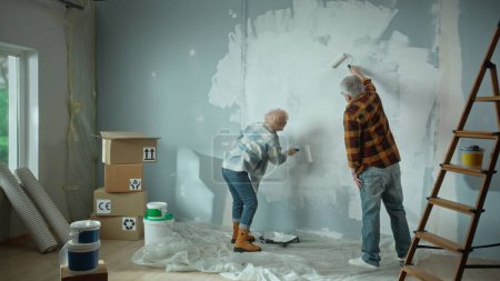 Photo for Elderly man and woman are painting wall with white paint using paint rollers. Couple of pensioners are making repairs to their apartment, in the background of window, stepladder, cardboard boxes - Royalty Free Image