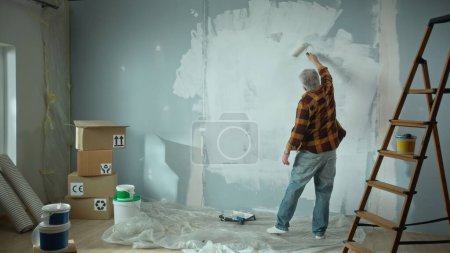 Photo for Elderly man is painting wall with white paint using paint roller. Pensioner is making repairs to their apartment, in the background of a window, stepladder, cardboard boxes, wallpaper and paint. Back - Royalty Free Image