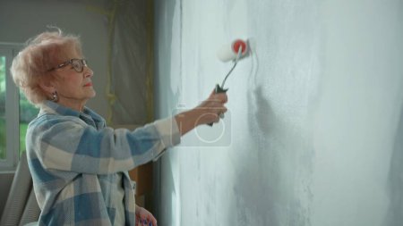 Foto de Mujer anciana está pintando la pared con pintura blanca usando rodillo de pintura. La pensionista está haciendo reparaciones en su apartamento con el telón de fondo de una ventana con luz solar brillante. Vista lateral. Concepto de - Imagen libre de derechos