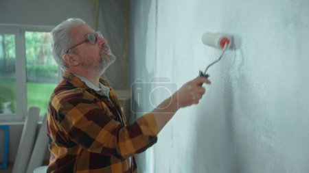 Photo for Elderly man is painting wall with white paint using paint roller. Male pensioner is making repairs to his apartment against the backdrop of a window with bright sunlight. Side view. Concept of repair - Royalty Free Image