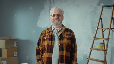 Téléchargez les photos : Elderly gray haired man with beard in glasses is looking at camera and smiling. Portrait of pensioner in plaid shirt is posing in an apartment against the background of a wall painted with white paint - en image libre de droit