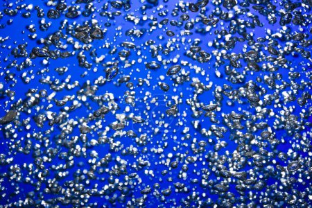 Foto de Una gran cantidad de burbujas de aire brillantes de diferentes tamaños bajo el agua sobre un fondo azul. El primer plano de burbujas de oxígeno iluminadas por la luz fluye hacia arriba. Aireación o filtración de líquido. Fizzy corriente de burbujas de aire - Imagen libre de derechos