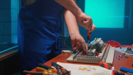 Foto de Unrecognizable man in blue overalls with a screwdriver in his hands is screwing a red wire in an automatic electrical switch. Electrical workshop with tools on the table, blue light - Imagen libre de derechos