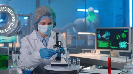 Photo for Modern medical research laboratory. Woman researcher in a white gown, mask, blue gloves and a bonnet is examining a sample in a microscope. People in white hazmat suits are working in the background - Royalty Free Image