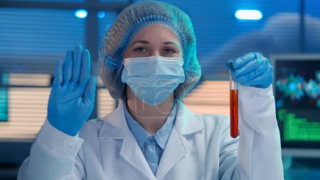 Téléchargez les photos : Une femme scientifique tient une éprouvette avec un liquide rouge à la main et montre un geste d'arrêt avec sa paume. Une femme en gants bleus, une blouse médicale blanche et un masque montrent un geste d'avertissement. Gros plan - en image libre de droit