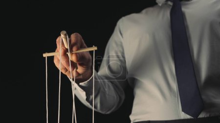 Téléchargez les photos : Un homme d'affaires en chemise grise et cravate noire contrôle une marionnette avec un manipulateur en bois et des cordes. Un marionnettiste contrôle une marionnette sur un fond noir isolé. Le concept de contrôle - en image libre de droit