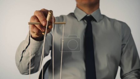 Téléchargez les photos : Un homme d'affaires en chemise grise et cravate noire contrôle une marionnette avec un manipulateur en bois et des cordes. Un marionnettiste contrôle une marionnette sur un fond blanc isolé. Le concept de contrôle - en image libre de droit