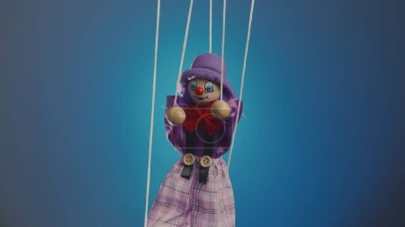 Foto de Payaso marioneta colgando de cuerdas. Muñeca de trapo con traje y sombrero púrpura, con lazo rojo, nariz y maquillaje. Muñeca suave arlequín funster en un fondo de estudio azul. El concepto de unas vacaciones para niños - Imagen libre de derechos