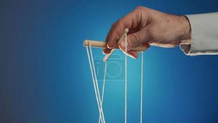 Photo for The hand of a man in a gray shirt controls a puppet using a wooden manipulator and strings on a blue background. Hand handling at puppet by pulling strings to make the character move. The concept of - Royalty Free Image