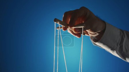 Téléchargez les photos : The hand of a man in a gray shirt controls a puppet using a wooden manipulator and strings on a blue background. Hand handling at puppet by pulling strings to make the character move. The concept of - en image libre de droit