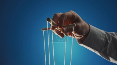 Photo for The hand of a man in a gray shirt controls a puppet using a wooden manipulator and strings on a blue background. Hand handling at puppet by pulling strings to make the character move. The concept of - Royalty Free Image