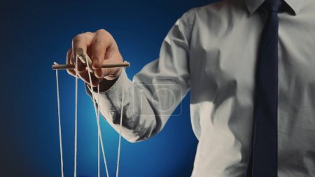 Photo for A businessman in a gray shirt and black tie controls a puppet with a wooden manipulator and strings. Hand handling at puppet by pulling strings to make the character move. Blue isolated background - Royalty Free Image