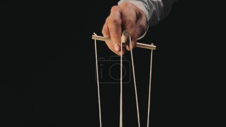 Téléchargez les photos : The puppeteers hand controls the puppet with a wooden manipulator and strings. The marionettist controls and pulls the strings on a black background. The concept of dependence, dominance, managing - en image libre de droit
