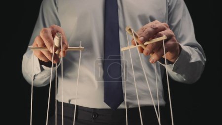 Téléchargez les photos : Un homme d'affaires en chemise grise et cravate noire contrôle une marionnette avec un manipulateur en bois et des cordes. Le marionnettiste manipule la marionnette en tirant les cordes avec les deux mains sur un fond noir - en image libre de droit