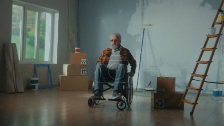 Téléchargez les photos : Un handicapé âgé en fauteuil roulant. Le pensionné regarde vers l'avenir et planifie des réparations. Chambre avec fenêtre, échelle, boîtes en carton, rouleaux de papier peint, rouleau et peinture. Concept de réparation dans l'appartement - en image libre de droit