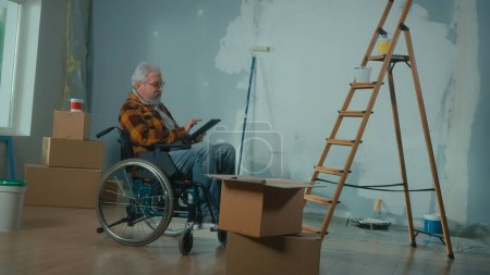 Foto de Un anciano discapacitado se mueve en una silla de ruedas planea la renovación usando una tableta digital. Habitación con ventana, escalera, cajas de cartón, rollos de papel pintado, cubo de pintura. Concepto de reparación en el apartamento. - Imagen libre de derechos