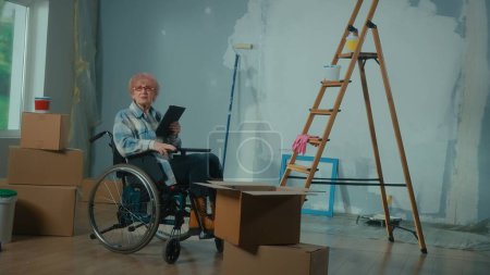 Photo for An elderly disabled woman in a wheelchair plans renovation using a digital tablet. Room with window, ladder, cardboard boxes, wallpaper rolls, paint bucket. Concept of repair in the apartment. - Royalty Free Image