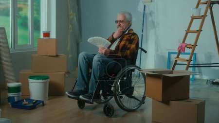 Foto de Un anciano discapacitado en silla de ruedas mira a través de la paleta con colores. Un pensionista planea reparaciones y elige un color de pintura. Habitación con ventana, escalera, cajas de cartón, rollos de papel pintado, pintura - Imagen libre de derechos