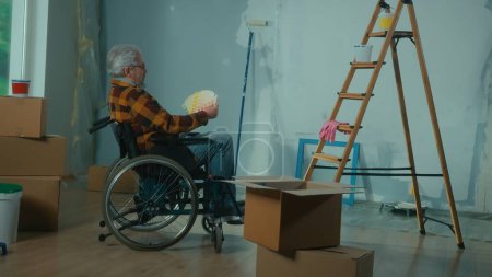 Téléchargez les photos : Un handicapé âgé en fauteuil roulant regarde à travers la palette avec des couleurs. Un pensionné planifie des réparations et choisit une couleur de peinture. Chambre avec fenêtre, échelle, boîtes en carton, rouleaux de papier peint, peinture - en image libre de droit