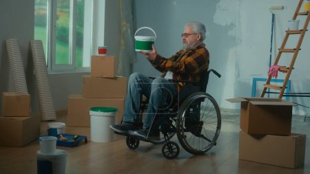 Téléchargez les photos : Un homme handicapé âgé en fauteuil roulant examine un seau de peinture. Un pensionné planifie des réparations et une décoration murale. Chambre avec fenêtre, échelle, boîtes en carton, rouleaux de papier peint. Concept de réparation dans le - en image libre de droit