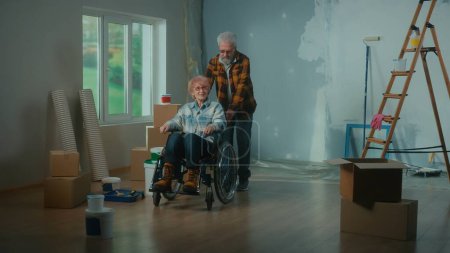 Téléchargez les photos : Un homme âgé et une femme âgée en fauteuil roulant. Un couple de personnes âgées planifie des réparations sur fond de fenêtre, d'échelle, de boîtes en carton, de seaux de peinture, de rouleaux et de brosses. Le concept - en image libre de droit
