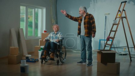 Téléchargez les photos : Un homme âgé et une femme âgée en fauteuil roulant. Un couple de personnes âgées communique et planifie des réparations. Chambre avec une fenêtre, échelle, boîtes en carton, seaux de peinture, rouleaux et brosses. Le concept - en image libre de droit