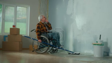 Foto de Un anciano en silla de ruedas pinta una pared con pintura blanca usando un rodillo largo. Un hombre discapacitado en la edad hace reparaciones en la habitación. El concepto de reparación en el apartamento realizado por personas con - Imagen libre de derechos