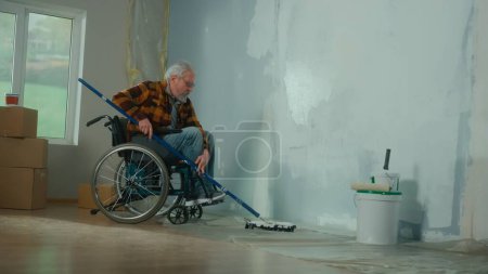 Photo for An elderly man in a wheelchair paints a wall with white paint using a long roller. A disabled man in the age makes repairs in the room. The concept of repair in the apartment performed by people with - Royalty Free Image