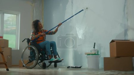 Foto de Una mujer afroamericana en silla de ruedas pinta una pared con pintura blanca usando un rodillo largo. Una joven negra con discapacidad hace reparaciones en la habitación y sonríe. El concepto de renovación en el - Imagen libre de derechos