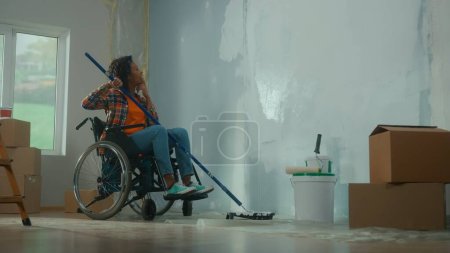 Foto de Una mujer afroamericana en silla de ruedas con un rodillo largo en las manos mira cuidadosamente a la pared y planea pintar. Una joven negra con discapacidad hace reparaciones en la habitación. El concepto de - Imagen libre de derechos
