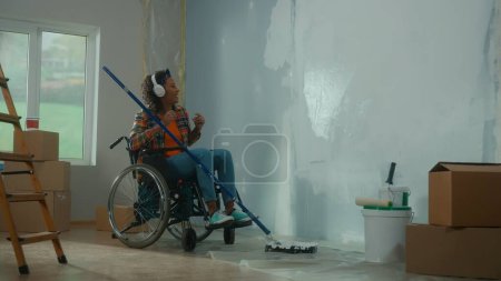 Foto de Una mujer afroamericana con auriculares blancos en silla de ruedas pinta una pared con pintura blanca usando un rodillo largo. Una joven negra con discapacidad hace reparaciones en la habitación y disfruta de la música. El - Imagen libre de derechos