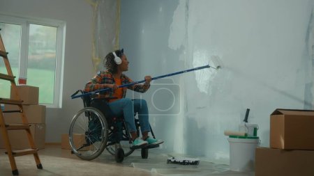 Téléchargez les photos : Une Afro-Américaine en casque blanc sur un fauteuil roulant peint un mur avec de la peinture blanche à l'aide d'un long rouleau. Une jeune femme noire handicapée fait des réparations dans la chambre et aime la musique. L ' - en image libre de droit