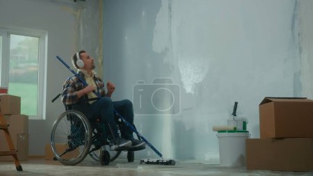 Téléchargez les photos : Un jeune homme en casque blanc sur un fauteuil roulant prévoit de peindre un mur avec de la peinture blanche à l'aide d'un long rouleau. Un handicapé fait des réparations dans la pièce et aime la musique. Le concept de rénovation dans le - en image libre de droit