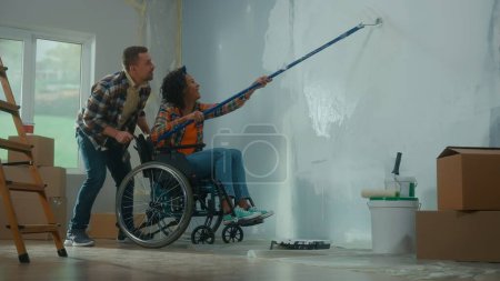 Téléchargez les photos : An African American woman in a wheelchair paints a wall with white paint using a long roller. A black woman and a white man make repairs in the room and laugh happily. A young couple. The concept of - en image libre de droit