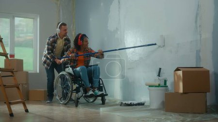 Foto de Un hombre blanco y una mujer afroamericana en silla de ruedas hacen reparaciones en la habitación y disfrutan de la música con auriculares. Una joven pareja pinta una pared con un rodillo largo y sonríe felizmente. El concepto de - Imagen libre de derechos