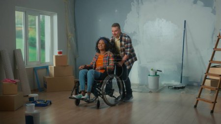 Foto de Un hombre blanco y una mujer afroamericana en silla de ruedas. Una pareja joven está planeando una reparación en la habitación. Una habitación con una ventana, una escalera, cajas de cartón, rollos de papel pintado y cubos de pintura. El - Imagen libre de derechos