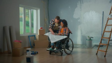 Téléchargez les photos : A white man and an African American disabled woman in a wheelchair are looking at an apartment project sheet. A young couple is planning a renovation in the room, talking and discussing interior - en image libre de droit