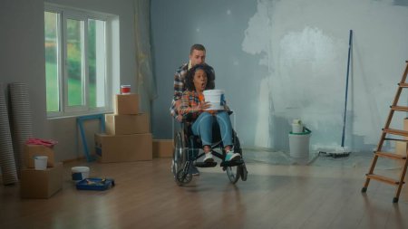 Téléchargez les photos : Un homme blanc et une femme afro-américaine en fauteuil roulant avec un seau de peinture dans les mains. Femme noire avec un visage surpris et une émotion choquée. Le concept de rénovation dans l'appartement - en image libre de droit