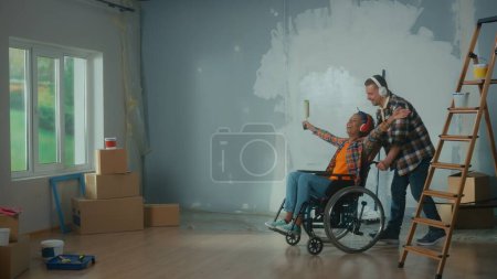 Téléchargez les photos : Un homme blanc conduisant une Afro-Américaine en fauteuil roulant dans la pièce. Un jeune couple dans de grands écouteurs aime la musique pendant la rénovation. Le concept de rénovation dans l'appartement. - en image libre de droit