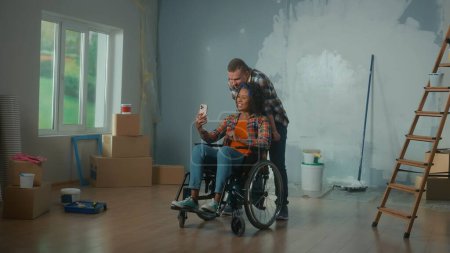 Foto de Un hombre blanco y una mujer afroamericana discapacitada en silla de ruedas están hablando en videollamada usando un teléfono móvil. Una pareja joven comunica y comparte planes para reparar la habitación. El concepto de - Imagen libre de derechos
