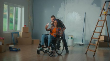 Téléchargez les photos : Un homme blanc et une femme handicapée afro-américaine en fauteuil roulant parlent sur appel vidéo à l'aide d'un téléphone portable. Un jeune couple communique et partage des plans pour réparer la pièce. Le concept de - en image libre de droit