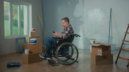 Téléchargez les photos : Un jeune homme en fauteuil roulant planifie des réparations à l'aide d'un téléphone mobile. Handicapés masculins navigation idées de design d'intérieur sur Internet. Handicapés sur le fond des boîtes en carton et des fenêtres. L ' - en image libre de droit
