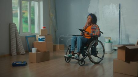 Téléchargez les photos : Young African American woman in a wheelchair is planning a renovation using a digital tablet. Disabled woman browses interior design ideas on the internet. Handicapped person on the background of - en image libre de droit