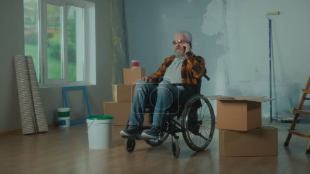 Téléchargez les photos : Un handicapé âgé en fauteuil roulant parle sur un téléphone portable. Personne retraitée handicapée. Chambre avec fenêtre, boîtes en carton, rouleaux de papier peint, seau à peinture. Le concept de rénovation dans le - en image libre de droit