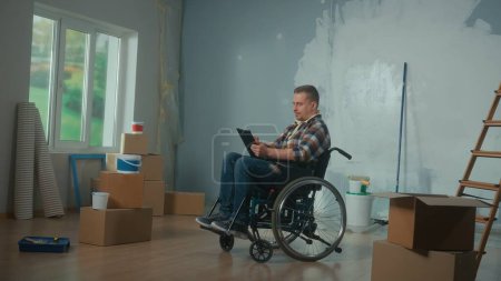 Téléchargez les photos : Un jeune homme en fauteuil roulant planifie des réparations à l'aide d'une tablette numérique. Handicapés masculins navigation idées de design d'intérieur sur Internet. Handicapés sur le fond des boîtes en carton et des seaux de - en image libre de droit