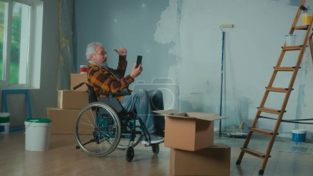 Foto de Un anciano discapacitado en silla de ruedas habla en una videollamada usando un teléfono móvil. Un pensionista macho apunta a la ventana. Una habitación con ventana, rollos de papel pintado, cajas de cartón y un cubo de pintura - Imagen libre de derechos