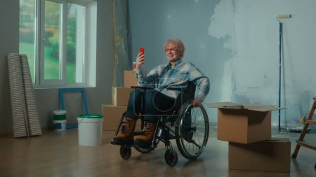 Téléchargez les photos : Une femme handicapée âgée en fauteuil roulant parle lors d'un appel vidéo à l'aide d'un téléphone mobile. Une pièce avec une fenêtre, une échelle, du papier peint, des boîtes en carton et un seau de peinture. Concept de réparation dans l'appartement - en image libre de droit