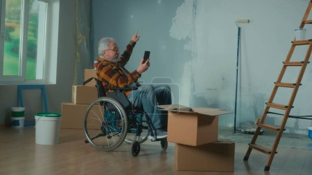 Photo for An elderly disabled man in a wheelchair talks on a video call using a mobile phone. A pensioner male points his finger at the wall. A room with window, wallpaper rolls, cardboard boxes and a bucket of - Royalty Free Image