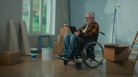 Téléchargez les photos : Un homme handicapé âgé déménage dans un fauteuil roulant plans de rénovation à l'aide d'une tablette numérique. Chambre avec fenêtre, échelle, boîtes en carton, rouleaux de papier peint, seau à peinture. Concept de réparation dans l'appartement. - en image libre de droit