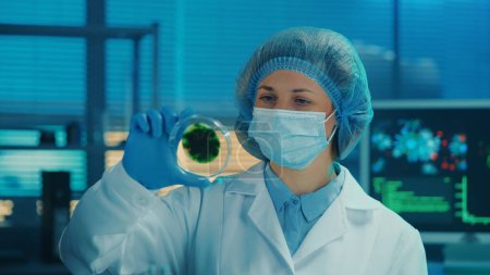 Photo for A female scientist examines a petri dish with a spotted green sample. A woman in a white medical gown, mask, blue gloves and a bonnet examines the spores of a virus or bacteria. A researcher works in - Royalty Free Image